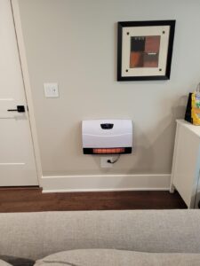Wall-mounted Heat Storm infrared heater installed above an outlet in a living room, glowing orange through the lower grille.