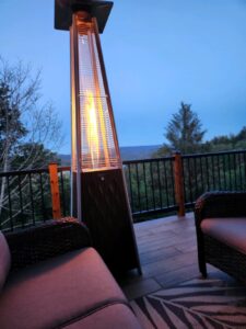 Tall pyramid patio heater lit on a deck at twilight, placed beside outdoor lounge chairs with a mountain view