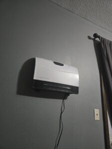 Heat Storm wall-mounted heater shown on a textured gray wall near a window curtain, side angle with power cord hanging down.