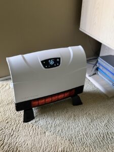 Heat Storm infrared heater with floor feet on carpet, digital display lit and heating element glowing, placed beside a desk.