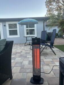 Infrared tower patio heater glowing red on a paver patio near lounge chairs and an umbrella table, plugged in for outdoor use.