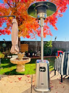Stainless patio heater on a backyard patio in autumn with red leaves, umbrella, and birdbath in the background