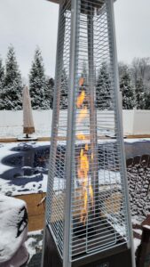 Pyramid patio heater burning in a snowy backyard by the pool, tall glass tube flame behind protective grille