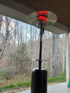 Lit propane patio heater under a covered patio, red heat glow visible beneath the reflector with trees in the background.