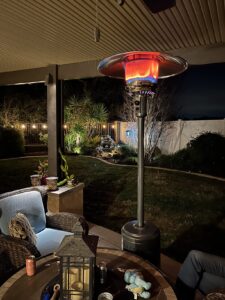 Outdoor propane patio heater lit on a covered patio beside seating area, warming a backyard at night